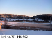 Winter rural landscape at sunset. The sun illuminates the snow-covered forest along the horizon, creating a warm golden-pink hue on the tops of the fir trees. In the foreground, a snow-covered slope with a built snowman and part of the roof of a house is visible. Below, a frozen river or lake, covered with thin ice and snow, with dry reeds growing around it, stretches out. The calm atmosphere of solitude, a cold winter evening and the beauty of nature is emphasized by the soft light of the fading day and a clear blue sky. Стоковое фото, фотограф Анна Кривицкая / Фотобанк Лори