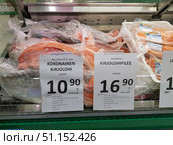 A supermarket counter with chilled packaged fish. In the foreground are large pieces and whole rainbow trout (kirjolohi) wrapped in film. Above the fish are price tags: on the left is the price for a whole fish from Finland - €10.90/kg, on the right is the price for a rainbow trout fillet from Sweden - €16.90/kg. In the background are plastic trays with packaged fillets. The image illustrates the selection of fresh fish in the sales area, the contrast in prices and product cutting options. (2019 год). Редакционное фото, фотограф Анна Кривицкая / Фотобанк Лори