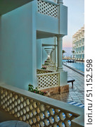 Architectural corridor overlooking the pool and the sea. White columns and decorative lattices create the effect of deep perspective and rhythm, stretching into the distance. Flower pots are visible between the columns, adding bright accents. On the left there is a patterned wall, on the right - a path by the water. On the horizon you can see palm trees and a calm sky in pastel shades of sunset. The atmosphere of coziness and relaxation emphasizes the resort mood, perfectly conveying the style of a luxury hotel by the sea. Стоковое фото, фотограф Анна Кривицкая / Фотобанк Лори