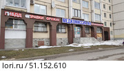 Facade of a multi-story residential building with commercial premises located on the ground floor. The building has bright signs of organizations and services: "Traffic Police", "Certificates", "Weapons", "Medical Commission No. 1", "Medical Examinations". A porch with stairs and railings leading to the entrance is visible. The windows are protected by metal bars. The lower part of the facade is made of gray brick and stone cladding, damage and cracks are visible on it. There are still remnants of snow on the street, and patches of green grass are visible near the sidewalk. The atmosphere is typical of the urban environment of Russia with the characteristic architecture of panel houses and a variety of commercial advertisements. (2019 год). Редакционное фото, фотограф Анна Кривицкая / Фотобанк Лори