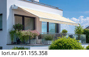 Modern balcony with striped retractable awning, lush plants, and glass railing on a sunny day, attached to a white contemporary residential building. Стоковое AI-изображение, AI-художник Катерина Бондарец / Фотобанк Лори