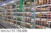 Supermarket display cases with a large selection of dairy products. Refrigerated shelves are filled with yoghurts, curd desserts and organic products of various brands in plastic and cardboard cups. Packaging of different colors - white, green, orange, pink, purple - create a bright and attractive visual series. Advertising stickers and price tags are placed on the glass doors of the refrigerator. Bright green and yellow signs with inscriptions are noticeable in the foreground. The atmosphere of a typical grocery store, an emphasis on the abundance of choice and modern design of the sales area. Редакционное фото, фотограф Анна Кривицкая / Фотобанк Лори