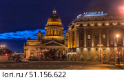A night photo of St. Petersburg with a view of St. Isaac's Cathedral and the Astoria Hotel. The frame is filled with the atmosphere of an evening city: the majestic dome of the cathedral is illuminated with golden lights, emphasizing the architectural beauty and scale of the building. Nearby is the facade of the historic Astoria Hotel with a bright neon sign reflecting the rich history and status of this place. In the foreground is a deserted street with rare cars and traffic lights, adding dynamics and creating a sense of calm and comfort of the night city. The deep blue color of the sky and the soft lighting of the lanterns create a harmonious combination and emphasize the romantic atmosphere of St. Petersburg. (2020 год). Редакционное фото, фотограф Анна Кривицкая / Фотобанк Лори