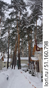 Winter village in a pine forest. A narrow path, cleared of snow, leads past private houses with white roofs covered with a thick layer of snow. Snowdrifts are visible on both sides of the road, and tall pine trees with reddish-brown trunks and fluffy crowns covered with snow are all around. On the right in the foreground is a house with a porch and a canopy, on the left you can see other houses with windows. The atmosphere is calm and peaceful, emphasizing the beauty of a winter forest village and the comfort of country life. Стоковое фото, фотограф Анна Кривицкая / Фотобанк Лори