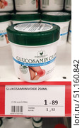 Jar of Herbamedicus Glucosamin Creme glucosamine skin cream displayed on a store shelf. The packaging is white with a green lid. The label shows the product name, the manufacturer's logo, and an image of a knee, symbolizing the use of the cream to relieve joint pain. Below the jar, on the price tag, the name in Finnish is "Glukosamiinivoide 250 ml", the price is 1.89 euros, and the cost without tax is 1.52 euros. In the background, other jars of the same product are visible, lined up in a row. (2019 год). Редакционное фото, фотограф Анна Кривицкая / Фотобанк Лори