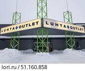 Facade of the Luhta Outlet shopping center in winter. In the foreground, a large illuminated sign with the LUHTA brand logo is visible, attached to green metal structures. Under the sign is the entrance to the store, surrounded by snowdrifts and a cleared path. A layer of snow and icicles are visible on the roof, which emphasizes the cold northern climate. The store is located in a modern gray building, the design of which is minimalist and functional. The atmosphere of the photo conveys the reality of everyday shopping in a snowy winter. (2019 год). Редакционное фото, фотограф Анна Кривицкая / Фотобанк Лори
