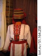 Traditional national women's costume, presented on a mannequin. The costume is made in white with red decorative elements and geometric embroidery. The decoration is red and gold patterns using traditional motifs. A wide woven belt with tassels is tied around the waist, complementing the image. On the head is a high headdress, richly decorated with multi-colored embroidery and gold trim. This costume reflects the ethnic heritage and originality of folk art, often used in ethnographic museums, exhibitions, folk culture festivals and traditional holidays. Стоковое фото, фотограф Анна Кривицкая / Фотобанк Лори