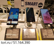 Electronics store windows with modern Samsung smartphones on display. The image shows the Samsung Galaxy A9 (2018), Samsung Galaxy S9, and Samsung Galaxy S9 Plus. The smartphones are mounted on stands with protective alarm systems and have bright screens with different screensavers. Under each device are price tags with specifications and prices: Galaxy A9 — €549, Galaxy S9 — €399, Galaxy S9 Plus — €599. It is clear that the information on the price tags is written in Finnish, indicating that the shooting took place in a Finnish store. Above the smartphones is an advertising poster with the inscription SMART and a mention of the game Fortnite, available on Samsung devices. (2019 год). Редакционное фото, фотограф Анна Кривицкая / Фотобанк Лори