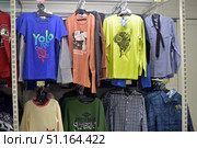 Rack in a clothing store with men's and teenage clothes hanging on it. The hangers contain long-sleeved T-shirts, shirts, and long sleeves of different colors: a blue T-shirt with a bright inscription, an orange long sleeve with a print, a light green T-shirt with a graphic design, a light blue shirt with a tie, checkered shirts in blue and blue-black shades. Green and light models with various inscriptions and designs hang on the lower rows. The photo conveys the atmosphere of a casual store aimed at young people and teenagers. (2015 год). Редакционное фото, фотограф Анна Кривицкая / Фотобанк Лори