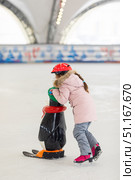 Side view little girl in warm pink coat and red safety helmet holding support and learning to skate on ice skating rink. Fall down and have fun. Winter sport, outdoors. Holiday and seasonal concept. Стоковое фото, фотограф Ирина Михайличенко / Фотобанк Лори