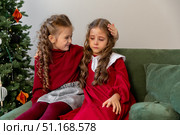 Two little girls sisters are sitting on a sofa near a Christmas tree. One girl is upset and cries, the other one hugs her and calms her down. Childhood and friendship relations concept. Стоковое фото, фотограф Ирина Михайличенко / Фотобанк Лори