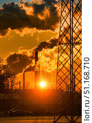 Industrial landscape at sunset. In the foreground is the metal structure of a power line, with part of its lattice tower to the right. On the horizon rise factory chimneys, from which rise thick smoke, tinted in the bright orange-yellow tones of the setting sun. The low sun creates a dramatic atmosphere, breaking through the clouds and plumes of smoke, giving the frame a spectacular combination of natural beauty and industrial influence. Below, silhouettes of cars and houses are visible, emphasizing the theme of urban life and ecology. Стоковое фото, фотограф Анна Кривицкая / Фотобанк Лори