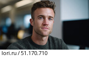 A confident young man sits in an office setting, looking directly at the camera with a subtle smile. His relaxed yet professional demeanor and casual sweater suggest a workplace environment that blends formality with approachability. The background includes blurred computer monitors, enhancing the corporate or tech-oriented atmosphere. This image fits themes related to modern workplace culture, professional identity, or digital careers, and can align with observances like Russia’s Day of the Programmer or Day of the Entrepreneur. Стоковое AI-изображение, AI-художник Ксения Чунаева / Фотобанк Лори