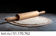 A wooden rolling pin resting on a piece of rolled-out dough placed on a stone countertop, set against a dark background. The scene evokes a rustic, homey kitchen atmosphere, ideal for baking or preparing traditional flatbreads, pizza, or pastries. Стоковое AI-изображение, AI-художник Ксения Чунаева / Фотобанк Лори