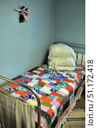 A small simple bed with a metal frame in a country room. On the bed lies a bright patchwork quilt with colorful geometric patterns. The pillow is covered with a white hand-knitted napkin, creating an atmosphere of coziness and traditional rural interior. The walls are painted blue, two amulet dolls in folk costumes hang on the wall. The atmosphere of the image conveys the simplicity, modesty and homely warmth of village life. Стоковое фото, фотограф Анна Кривицкая / Фотобанк Лори