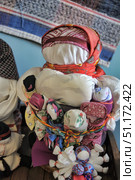 Close-up of a traditional folk amulet doll, handmade using the motanka technique. The doll is dressed in a bright scarf of red-orange shades and multi-layered clothing. The peculiarity of this doll is the many small dolls tied to its body, which symbolizes fertility, motherhood, care and family well-being. Rag dolls are made of fabric with various patterns and decorated with colored threads. The background is part of an ethnographic exhibition with a woven rug on the wall. The atmosphere of the photo emphasizes the connection with traditions, cultural heritage and the uniqueness of folk art. Стоковое фото, фотограф Анна Кривицкая / Фотобанк Лори