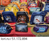 A bright showcase with handmade leather bags decorated with unique designer drawings. In the foreground are products with colorful images of characters - an owl with a cup of coffee, a white rabbit with a clock, the Cheshire cat from Alice in Wonderland, as well as stylized portraits. The bags are made in different colors: blue, burgundy, orange, green, brown and purple, which makes the composition rich and attractive. Each product looks exclusive, combining the traditions of craftsmanship with modern art design. Стоковое фото, фотограф Анна Кривицкая / Фотобанк Лори