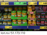 Supermarket shelves with canned vegetables. In the foreground are glass jars of pickled pumpkin from the Freshona and Felix brands. The products are neatly laid out in cardboard boxes with the company logos. In the background, other canned goods are visible: cucumbers, salads, and cans of Campolargo beans. Above the shelf are bright yellow and green price tags indicating the cost of the goods. The scene illustrates the atmosphere of a grocery store, the variety of products, and the design of the shelves with goods. (2015 год). Редакционное фото, фотограф Анна Кривицкая / Фотобанк Лори