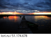 A picturesque sunset over a lake. In the foreground is an old wooden pier extending into the water, with a boat tied up nearby. The sky is covered with thick storm clouds, through which the bright orange-red glow of the setting sun breaks through. The reflections of the sunset and clouds create a spectacular play of light on the surface of the water. The atmosphere of the frame is both dramatic and calm, conveying the beauty of nature and the solitude of the moment. Ideal for themes of travel, nature, solitude, recreation on the water and philosophical images. Стоковое фото, фотограф Анна Кривицкая / Фотобанк Лори