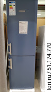Blue Liebherr refrigerator with a bottom freezer. The model is presented in the sales area, on the door you can see a label with the characteristics and price - 36,390 rubles. The refrigerator has a modern design with straight lines and two separate handles for the upper and lower doors. On the side there is a label with an energy class A++, which indicates high energy efficiency. The total volume of the refrigerator is 210 liters, of which the freezer takes up 84 liters. The color scheme in a sky-blue shade gives the equipment a stylish and unusual appearance, standing out against the background of standard white models. (2018 год). Редакционное фото, фотограф Анна Кривицкая / Фотобанк Лори