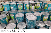 A photograph of supermarket shelves lined with cans of Heinz green peas. The easy-to-open metal cans come in signature green and turquoise packaging with the Heinz logo and the words "Tender Peas." The can lids with pull tabs are clearly visible in the foreground, and the labels with the ingredients and nutritional value of the product are in the background. The photo conveys an atmosphere of abundance and choice in the store, reflecting the theme of trade, consumption, and food packaging. (2021 год). Редакционное фото, фотограф Анна Кривицкая / Фотобанк Лори