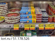 A refrigerated shelf in a store with an assortment of canned fish and seafood. On the left, large round tins of smoked eel in decorative packaging are visible. In the center are packages of Calvo brand canned tuna in blue boxes with the inscription Special Offer on yellow labels. Small plastic containers with red and black caviar are stacked nearby. On the right are glass jars with gold lids filled with red caviar. On the bottom shelf are packages of John West tuna and other canned fish. Under the products are price tags with bright markings Hea Hind (Good price), indicating promotional offers. The atmosphere of the photo conveys the appearance of an ordinary supermarket, where a wide selection of delicacies from fish and seafood is presented. (2015 год). Редакционное фото, фотограф Анна Кривицкая / Фотобанк Лори