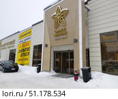 Facade of the Grande Orchidee fashion center in winter. In the foreground you can see the entrance with large glass doors and a large golden emblem in the form of an orchid, as well as the name of the center. On the left there is a large yellow banner with the inscription about the outlet closing and discounts from 30% to 80%, the text is duplicated in several languages ​​(Russian, Finnish). There is a black car in front of the building, snow lies on the ground, trash cans and a few snowdrifts are visible near the entrance. The atmosphere of the photo conveys a winter day at the entrance to a fashionable shopping center during a sale and liquidation of goods. (2019 год). Редакционное фото, фотограф Анна Кривицкая / Фотобанк Лори
