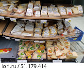 A supermarket display case with a large assortment of packaged cheeses. The cheeses are cut into pieces of various shapes and weights, each wrapped in transparent film and provided with a label with information about the manufacturer, weight and price. The upper and middle parts of the shelves contain more expensive varieties of cheese, mainly with white and beige shades, and at the bottom, pieces of Gouda cheese in branded packaging with green and yellow labels are clearly visible. The price tags under the products indicate the cost per kilogram, some products are highlighted with red stickers with a discount. The atmosphere of the photo emphasizes the variety of the assortment and the neat design of the shelves in the store. (2019 год). Редакционное фото, фотограф Анна Кривицкая / Фотобанк Лори