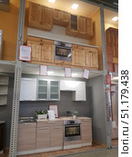Interior of a furniture store, where modular kitchen units are on display. The lower level shows a modern Scandinavian-style kitchen: the lower cabinets are made in a light beige wood shade with smooth facades, the upper cabinets are white glossy and with matte glass inserts. A hob and an oven made of stainless steel are built in. On the work surface there are decorative plants in pots, creating a cozy atmosphere.The upper part of the exhibition presents a more classic model of a kitchen made of natural wood with facades made of solid pine. The cabinets have a natural wood texture, which emphasizes environmental friendliness and traditional style. Both units are on display at the stand to demonstrate to customers possible kitchen design options. On the walls there are price tags with information about the cost and configuration. (2019 год). Редакционное фото, фотограф Анна Кривицкая / Фотобанк Лори