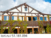 The facade of a timber-framed house. The white walls are divided by massive dark wooden beams, giving the building a traditional and rustic look. On the second floor, large windows with wooden frames and curtains are visible, reflecting the blue sky and clouds. The lower part of the facade is decorated with wicker elements made of vines, over which green and partly reddish wild grapes curl, creating the effect of a living wall. A decorative lantern is fixed to the pediment of the house, which adds color and completeness to the overall appearance. The atmosphere of the photograph conveys coziness, harmony with nature and traditional European architectural style.This image is ideal for illustrations on the topics of architecture, design of country houses, half-timbered buildings, country life, coziness and eco-friendly construction. Стоковое фото, фотограф Анна Кривицкая / Фотобанк Лори