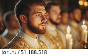 A young bearded man dressed in ornate ceremonial robes holds a lit candle during a solemn religious service. The soft glow of candlelight illuminates his face, reflecting a sense of reverence and spiritual devotion. Other men in similar attire appear blurred in the background, emphasizing the subject's contemplative expression. The image captures the peaceful and sacred atmosphere of a religious gathering or ritual. Стоковое AI-видео, аниматор Ксения Чунаева / Фотобанк Лори