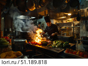Повар в черной униформе готовит на сковороде с огнем на кухне ресторана. Стоковое AI-изображение, AI-художник Кирилл Сиротюк / Фотобанк Лори