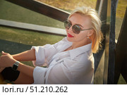 Young woman with blonde hair and sunglasses sits casually in warm sunlight, enjoying a serene moment outdoors. Стоковое фото, фотограф Кривошеина Елена Леонидовна / Фотобанк Лори