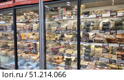 A supermarket display case with refrigerated shelves filled with a variety of ready-made and semi-finished products. Salads, appetizers, side dishes, meat and fish dishes, and desserts are laid out in plastic containers. Each package has price tags and stickers with information about the ingredients. The inscription “Main dishes for every taste” is noticeable at the top of the display case. Transparent glass doors of the refrigerators allow customers to easily examine the contents and choose the desired product. The scene emphasizes the modern format of supermarkets, which offer a wide range of ready-made food for fast eating and saving time. Редакционное фото, фотограф Анна Кривицкая / Фотобанк Лори