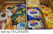 A shop window with various types of flatbreads and tortillas in factory packaging. On the right are neatly stacked packages of the Mission brand — classic flatbreads and cheese-flavored flatbreads in yellow and blue bags. On the left are products from other manufacturers: Mexican flatbreads, Armenian lavash, papadam (Indian crispy flatbreads), as well as various types of wheat and corn flatbreads. The packages are bright, colorful, and the inscriptions and logos of the brands are clearly visible. The photo conveys the atmosphere of a supermarket and demonstrates the range of bakery products for making shawarma, burritos, quesadillas, and other dishes. Редакционное фото, фотограф Анна Кривицкая / Фотобанк Лори