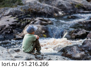 A little tourist boy is sitting with his back to the Akhinkoski Waterfall or the Ruskeala Waterfalls on the Tohmajoki River. Russia, Republic of Karelia. Стоковое фото, фотограф Photojuli / Фотобанк Лори