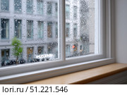 Raindrops sliding down window glass, blurring cityscape view with buildings and trees, evoking moody, contemplative urban rainy scene. Стоковое AI-изображение, AI-художник Юрий Поздников / Фотобанк Лори