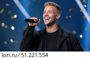 Male vocalist performing onstage, dressed in black, singing passionately with golden confetti falling, blue backdrop highlighting energetic performance. Стоковое AI-изображение, AI-художник Юрий Поздников / Фотобанк Лори