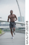 Athletic black man with a muscular physique is running shirtless, wearing black shorts and sneakers, on a paved path, with a modern bridge and city skyline in the background. Стоковое AI-видео, аниматор Юрий Поздников / Фотобанк Лори