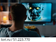 На темном фоне комнаты мужчина с миской чипсов смотрит на экран телевизора, где показано жуткое существо с горящими глазами. Стоковое AI-изображение, AI-художник Кирилл Сиротюк / Фотобанк Лори