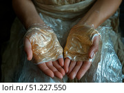 A close-up of hands wrapped in thin translucent films, creating an effect of fragility and vulnerability. The folds of the material shimmer with golden and silver shades, contrasting with the natural tone of the skin. The palms are open and facing upwards, symbolizing openness, trust or expectation. The background is made in soft tones, emphasizing the main focus on the texture of the film and the shape of the hands. The image can be interpreted in concepts of ecology, protection, art, modern society and the fragility of human nature. Стоковое AI-изображение, AI-художник Анна Кривицкая / Фотобанк Лори