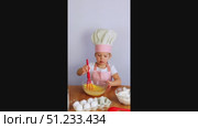 A little girl in a chef's hat and apron stirs dough in a bowl with a whisk. On the table are a eggs, flour, white background. Стоковое видео, видеограф Anna Scherbina / Фотобанк Лори