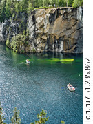 Marble Canyon in Ruskeala Mountain Park, Karelia. In the foreground is the deep blue water surface of the lake, sparkling in the sun. On the surface of the water are two boats with tourists: one closer to the cliff, the other at the bottom of the frame. A high sheer cliff with characteristic marble veins and shades of gray, white and brown rises above the lake, and from above you can see green trees and people standing on the observation deck. The water at the foot of the cliff is transparent, with green shades, through it you can see underwater formations. The atmosphere of the frame conveys the beauty of nature, tranquility and tourist appeal of this unique place. Стоковое фото, фотограф Анна Кривицкая / Фотобанк Лори