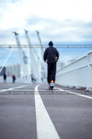 05/02/2025 Russia.Moscow.Person in dark clothing rides an electric scooter on a bridge pathway. Background includes blurred figures, tall structures, and a cloudy sky. Редакционное фото, фотограф Алина Лебедь / Фотобанк Лори