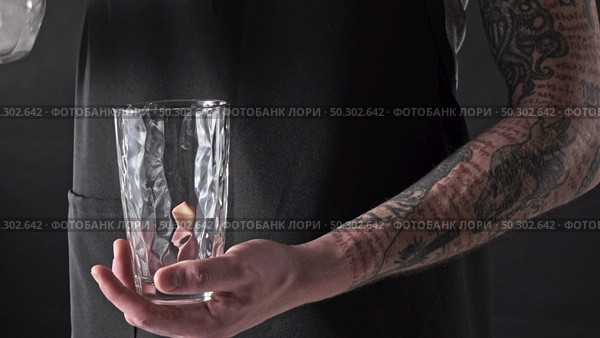A barista man pours ice cubes into a glass with splashes of drops on a dark background. 4K UHD video, 240fps, 2160p.. A man with a tattoo is pouring ice cubes into a glass around a dark background. 4K UHD video, 2160p. Стоковое фото, агентство Ingram Publishing / Фотобанк Лори