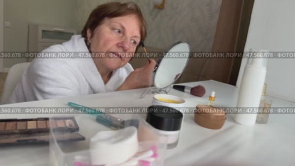A beautiful senior woman is sitting at home and doing her make-up morning routine. Senior woman putting blush on her face. Стоковое видео, видеограф Ирина Старикова / Фотобанк Лори