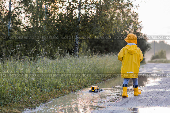 A boy in yellow rubber boots and a raincoat runs through puddles, having fun and playing with a remote-controlled car after it rains. Summer and autumn holidays. A happy childhood. Стоковое фото, фотограф Photojuli / Фотобанк Лори