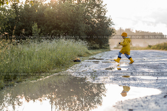 A boy in yellow rubber boots and a raincoat runs through puddles, having fun and playing with a remote-controlled car after it rains. Summer and autumn holidays. A happy childhood. Стоковое фото, фотограф Photojuli / Фотобанк Лори