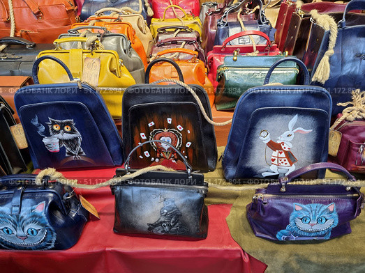 A bright showcase with handmade leather bags decorated with unique designer drawings. In the foreground are products with colorful images of characters - an owl with a cup of coffee, a white rabbit with a clock, the Cheshire cat from Alice in Wonderland, as well as stylized portraits. The bags are made in different colors: blue, burgundy, orange, green, brown and purple, which makes the composition rich and attractive. Each product looks exclusive, combining the traditions of craftsmanship with modern art design. Стоковое фото, фотограф Анна Кривицкая / Фотобанк Лори