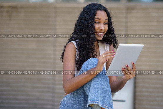 A cheerful and lively young woman is happily interacting with her tablet, effectively showcasing the significant role that technology plays in our daily lives, especially in an outdoor setting. A young woman is engaged with her tablet while sitting outdoors in a vibrant urban setting. Стоковое фото, агентство Ingram Publishing / Фотобанк Лори